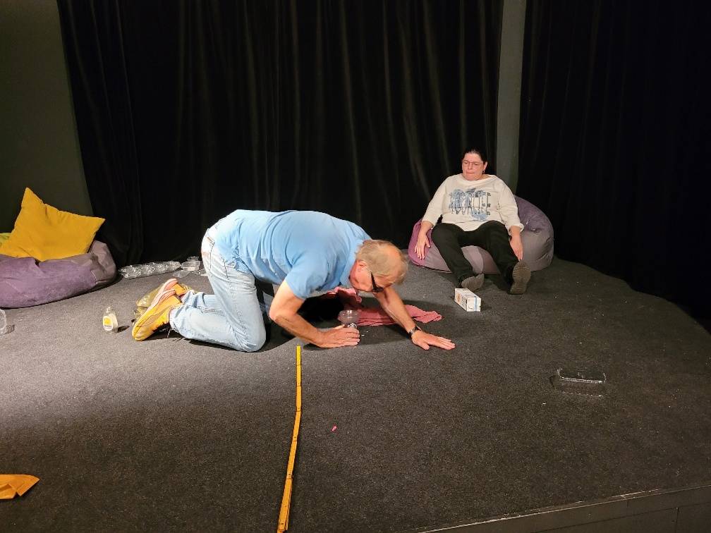 Eine Theaterszene: Eine Person kriecht auf allen Vieren über die Bühne, während eine andere entspannt auf einem Sitzsack sitzt und zusieht.