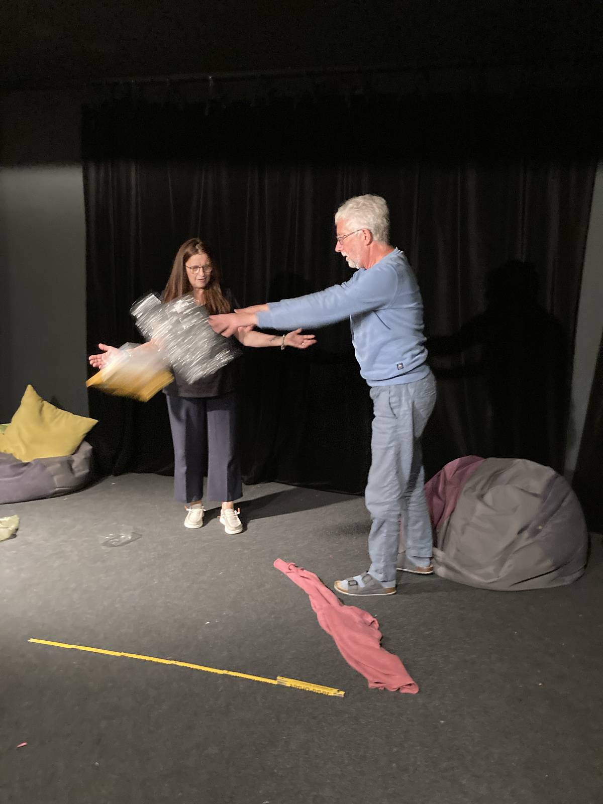 Eine Theaterszene: Zwei Personen auf der Bühne, eine wirft der anderen ein großes silbernes Objekt zu.