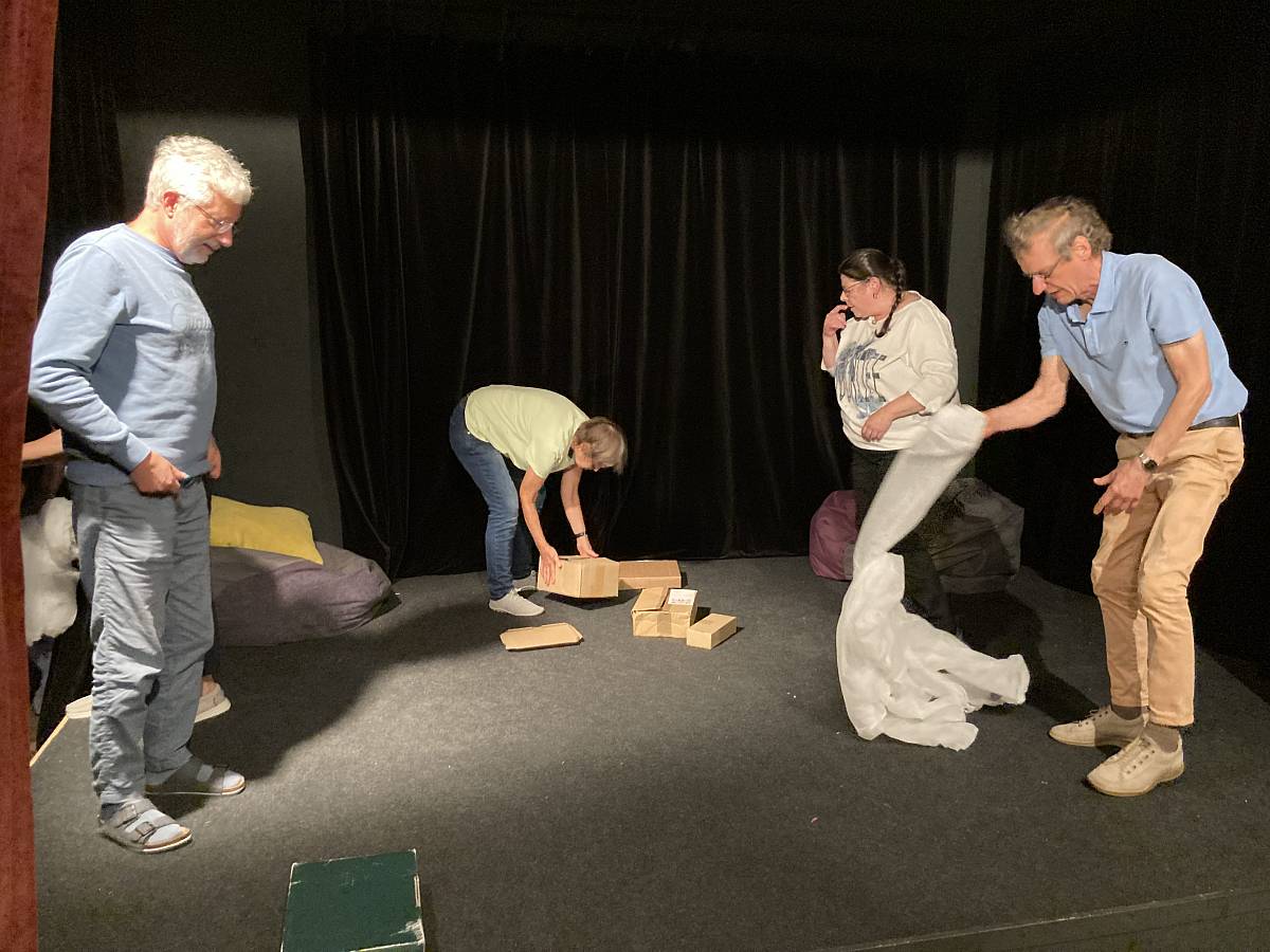 Vier Personen stehen und arbeiten auf einer Theaterbühne: Eine Person hebt Holzkisten auf, eine andere hält Stoff oder Folie, während die übrigen zuschauen oder sich bewegen.