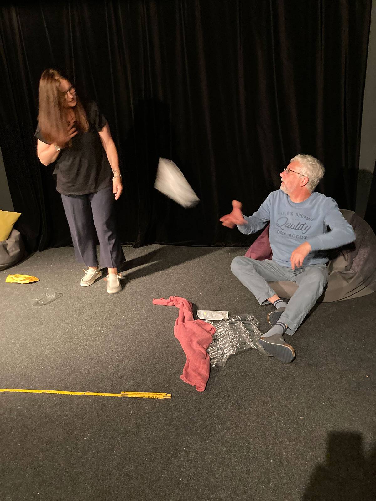 Eine Theaterszene: Eine Frau steht und schaut zu einem Mann, der auf einem Sitzsack sitzt und gerade ein Objekt in der Luft auffängt oder wirft.