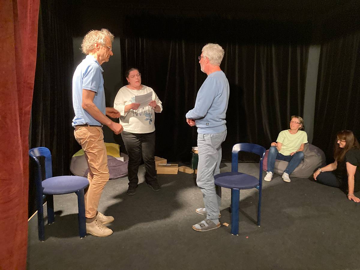 Fünf Personen auf einer kleinen Bühne mit schwarzem Vorhang; drei stehen und sprechen miteinander, eine Person hält ein Blatt Papier, während zwei weitere im Hintergrund auf Sitzsäcken sitzen.