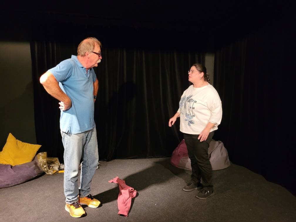 Zwei Personen stehen auf einer kleinen Bühne mit schwarzem Vorhang und sprechen miteinander; im Hintergrund liegen bunte Sitzkissen auf dem Boden.