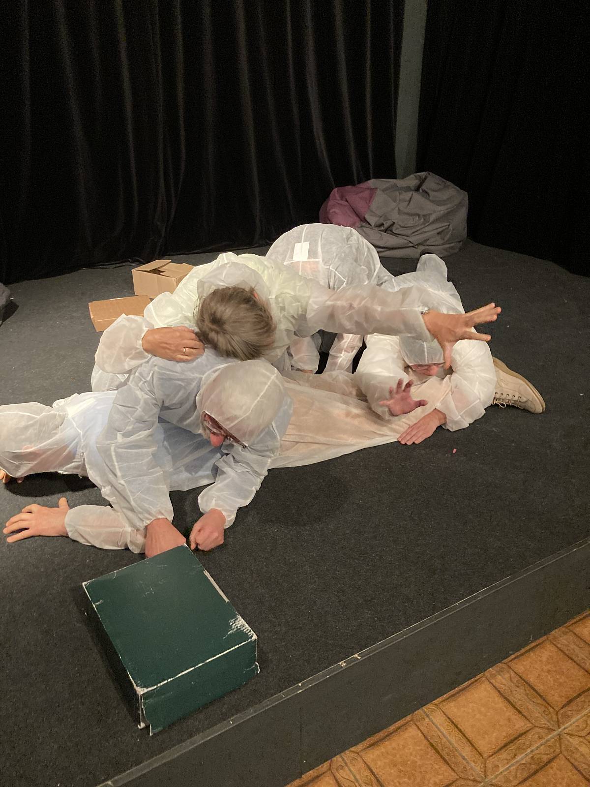 Vier Personen in weißen Overalls liegen und bewegen sich auf einer Theaterbühne in einer szenischen Darstellung.