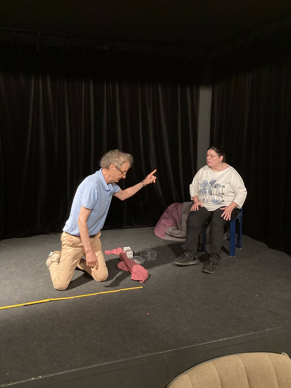 Zwei Personen auf einer kleinen Theaterbühne: Eine kniet auf dem Boden und gestikuliert mit erhobenem Finger, die andere sitzt auf einem Stuhl und schaut zu.